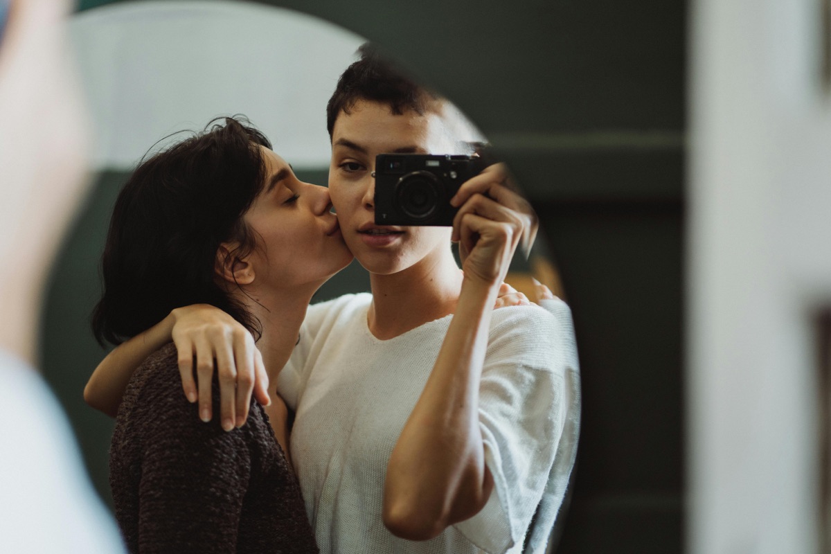 Theo and Ana in a mirror selfie, one kissing the other on the cheek while holding a camera.
